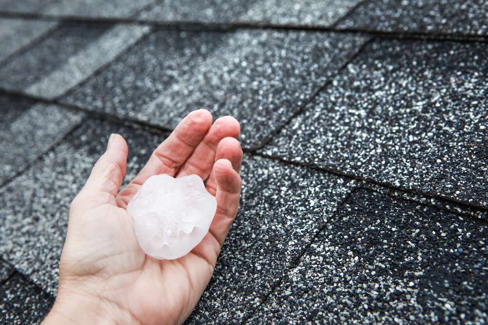 A hand holding a large hailstone above a roof.